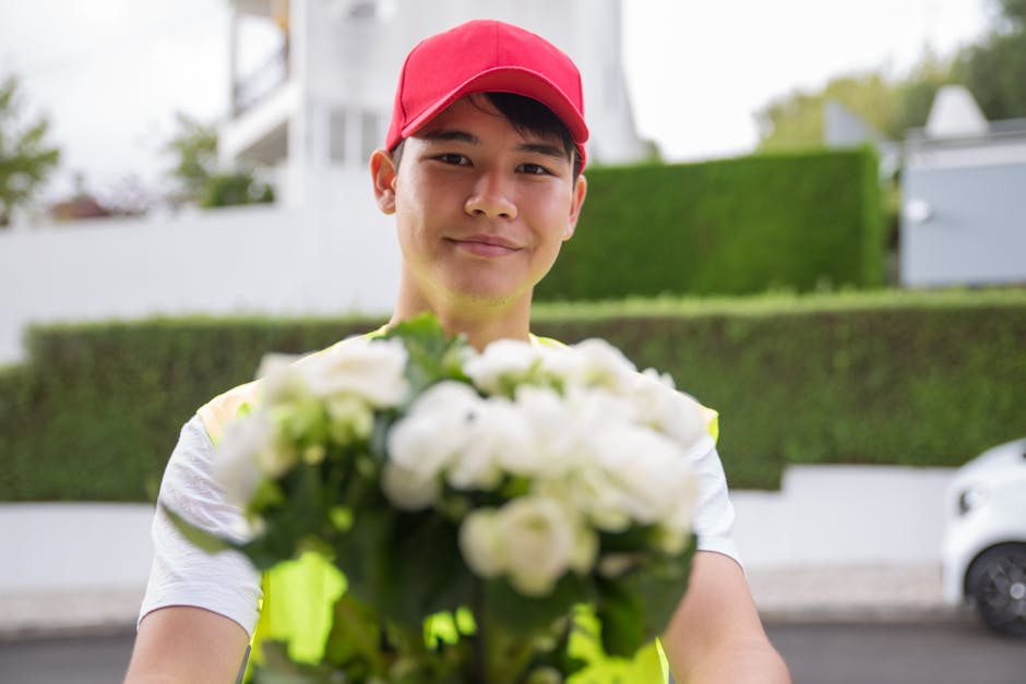 A young delivery person wearing a red cap and a high-visibility vest, smiling warmly while holding a bouquet of fresh white roses and greenery in front of them. The bouquet is arranged with a mix of f