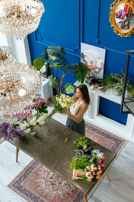 A woman arranging a vibrant bouquet of fresh flowers on a grey table inside a floral shop. The bouquet includes various greenery and white blossoms, with hints of pink and purple flowers visible. The