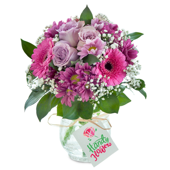 Centered in the frame and illuminated by clean, even lighting, this meticulously crafted floral bouquet captures the artistry and freshness synonymous with Northwood's reputable florists. Blossoming within the arrangement are soft lavender roses, their petals gracefully unfolding at the bouquet's heart to draw the eye. Nestled beside them, large fuchsia pink Gerbera daisies gleam, their dark-eyed centers expressing vivacity and cheer reminiscent of summer blooms along Northwood's local high streets. Smaller, plum-hued chrysanthemums with subtly golden-green centers create intricate textural layers, echoing the depth and diversity of Northwood's garden landscapes. Wisps of dainty white baby's breath float throughout, brightening the composition and instilling a gentle cloud-like softness. Enveloping the flowers are lush, deep green leaves, ensuring a harmonious natural framework. The bouquet sits in a striking clear vase, its 'cracked ice' glass glinting with rustic chic-an understated nod to contemporary tastes in the Northwood area. Around the vase's rim, a light brown raffia tie and a tidy white tag invite a handwritten note, ready to carry messages for occasions like birthdays, thank-yous, or heartfelt surprises. Every detail-down to the locally inspired flower selection-embodies the passion and expertise of Northwood florists, making this bouquet a truly memorable floral gift ready for prompt delivery to homes and workplaces in and around the neighbourhood.